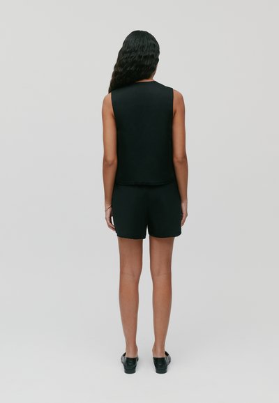 Zwart mouwloos topje en shorts met ruwe zoomdetails, gecombineerd met zwarte instappers, gezien vanaf de achterkant op een neutrale achtergrond.