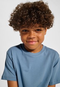 Barn med kort, lockigt brunt hår, klädd i en ljusblå T-shirt. Tröjan har en rund halsringning och en slät yta. Bakgrunden är enfärgad vit.