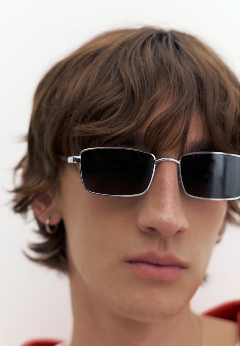 Jeune personne aux cheveux bruns ondulés portant des lunettes de soleil noires rectangulaires et un petit anneau à l’oreille, regardant devant elle sur un fond uni.