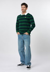 Άνδρας που στέκεται με τα χέρια στις τσέπες, φορώντας μακρυμάνικο πόλο με πράσινες και ναυτικές ρίγες, ανοιχτό μπλε τζιν και μαύρα αθλητικά παπούτσια σε λευκό φόντο.