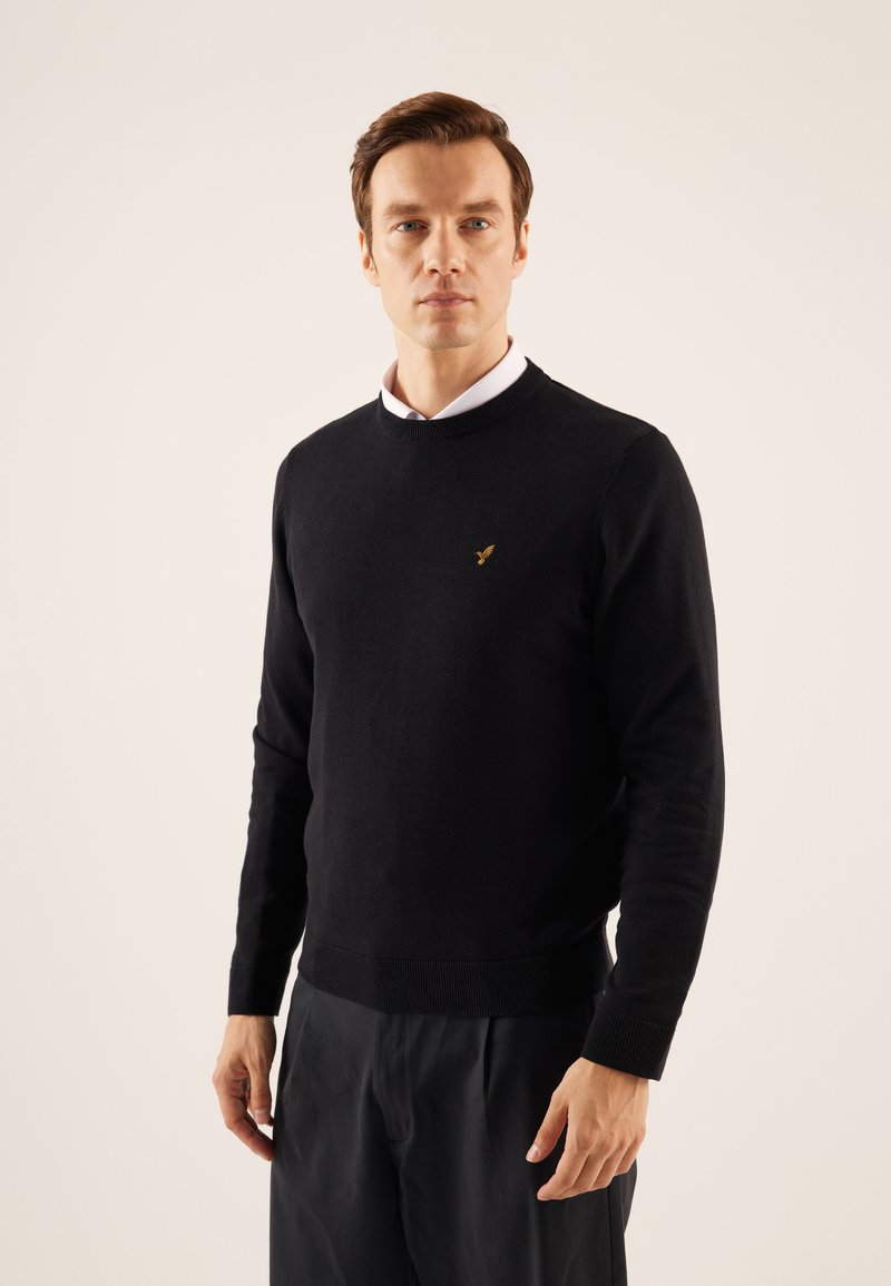 Pull en tricot noir à col rond, avec des poignets et un ourlet côtelés. Présente un petit logo doré sur la poitrine. Associé à un pantalon sombre.
