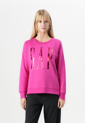 Fuchsia sweatshirt met "GAP" in reflecterende letters, ronde hals, lange mouwen en geribbelde manchetten; gedragen met zwarte broek.