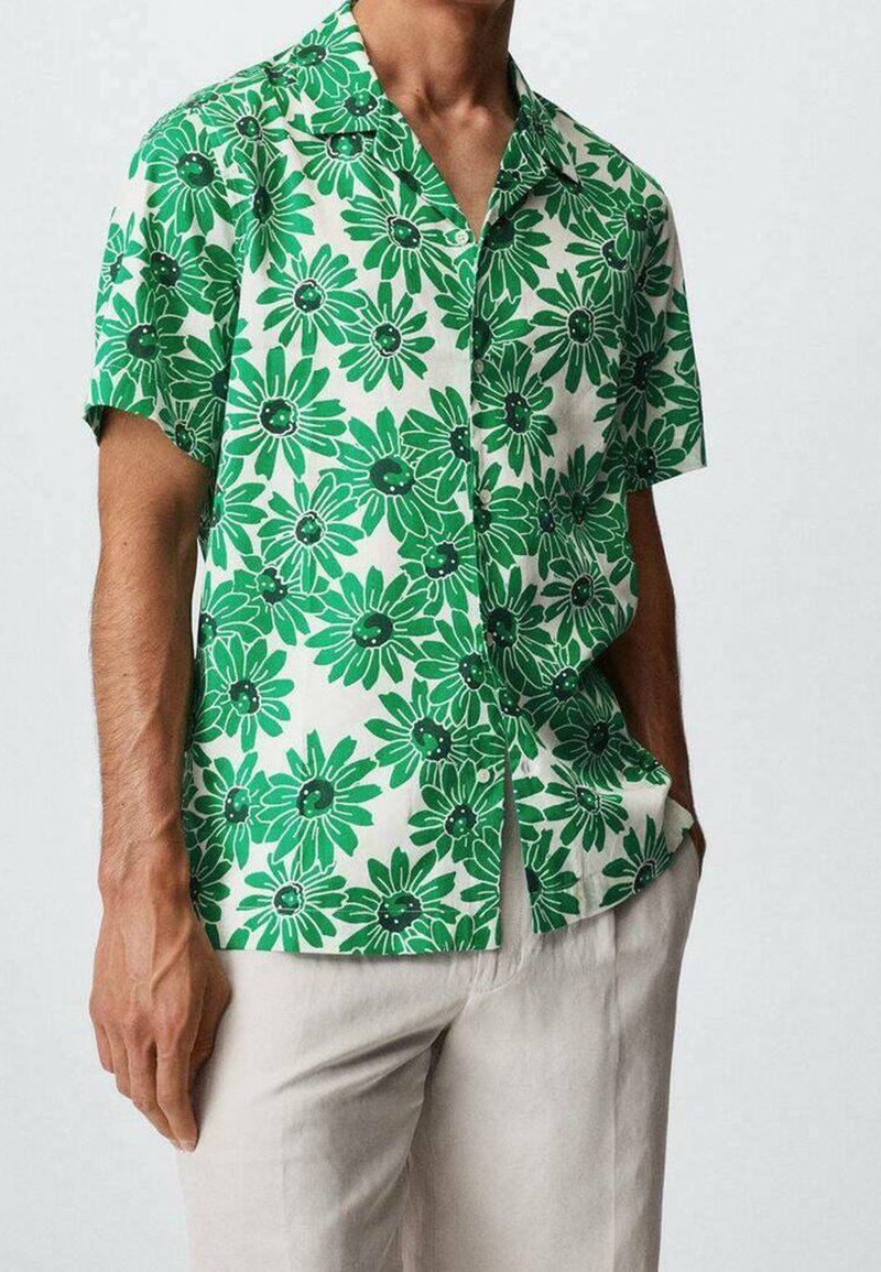 Homme portant une chemise à manches courtes avec un grand motif floral vert et un pantalon beige clair, une main dans la poche.