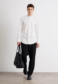 Camisa blanca de botones, pantalones negros y zapatos de cuero negros. Tote negra con un patrón recortado. Modelo caminando sobre una superficie clara.
