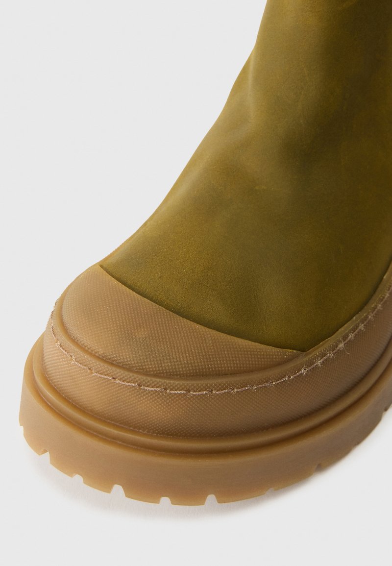 Olivgrüne Knöchelstiefel mit glattem Obermaterial, abgerundetem Zehenbereich und strukturiertem Gummi-Sohle. Verstärkte Nähte am Rand sorgen für Langlebigkeit.
