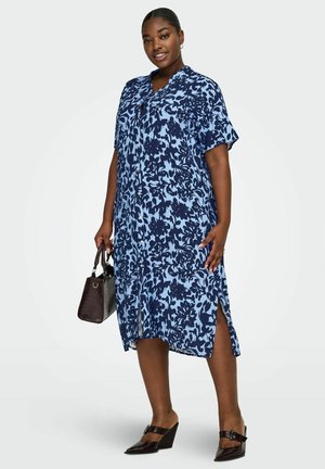 Kobieta w niebieskiej sukience midi w kwiaty z guzikami, trzymająca ciemną torebkę i nosząca czarne buty na obcasie, stojąca na białym tle.