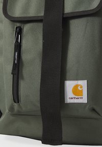 Grønn ryggsekk i stoffmateriale, med en frontlomme med glidelås, svart stropp og en hvit etikett med en oransje logo. Enkel, slitesterk design.