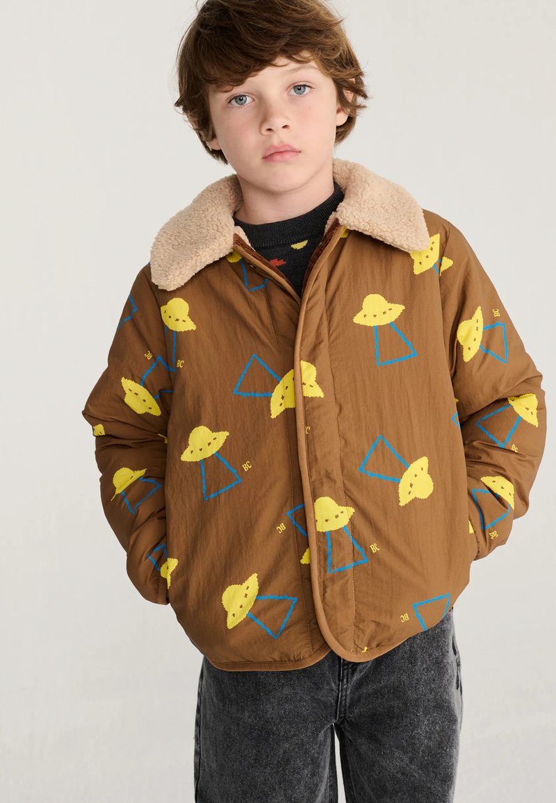 Garçon portant une veste marron avec des motifs OVNI jaunes et des triangles bleus et un col sherpa beige, debout les mains dans les poches.