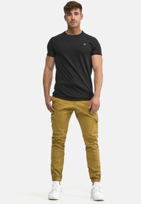 Tricou negru cu mâneci scurte și gât rotund, combinat cu pantaloni cargo muștar cu buzunare laterale și mansete înguste, completat cu adidași albi.