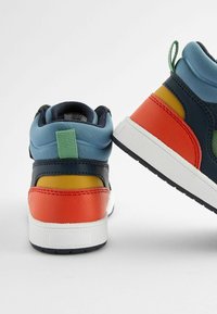 Wysokie sneakersy z niebieską cholewką, granatowymi skórzanymi akcentami, pomarańczowymi i żółtymi elementami, białą podeszwą oraz zielonym uchwytem z tyłu.