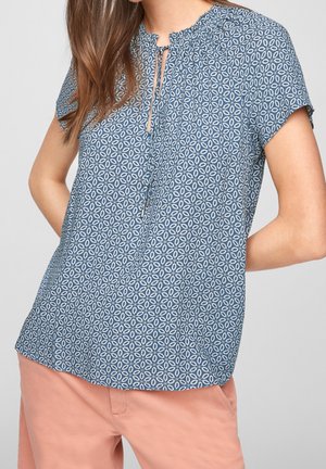 Vrouw draagt een blauw, kort mouwen blouse met geometrisch patroon en striksluiting bij de hals, gecombineerd met lichtroze broek, staand met handen achter haar rug.