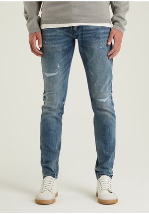 Hombre que lleva puestos unos jeans ajustados de mezclilla azules desgastados, zapatillas blancas con suelas de goma, y un suéter gris claro, de pie contra un fondo simple.