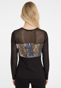 Zwarte langemouwen top met een transparante achterkant en een met pailletten bedekte paneel in de tinten zilver, goud en brons over het midden van de rug.