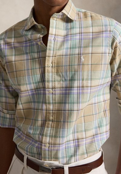 Homme portant une chemise à manches courtes à carreaux pastel, boutonnée, rentrée dans un pantalon beige avec une ceinture en cuir marron.