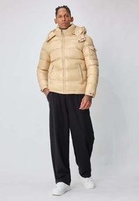 Homme debout, portant une doudoune beige avec capuche, pantalon ample noir, baskets blanches, sur fond uni gris clair.