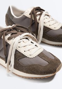 Paire de baskets marron et beige décontractées avec des panneaux en suède et tissu mélangés, dotée de lacets bicolores et de semelles en caoutchouc texturées.
