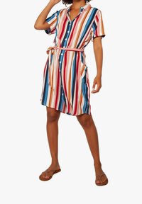 Donna che indossa un vestito camicia a righe verticali multicolori con una cintura in vita e infradito marroni, in piedi davanti a uno sfondo bianco.