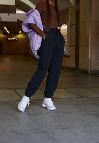 Osoba v černých teplákách, bílých teniskách a hnědém tričku, držící levandulovou prošívanou bundu vnitřním veřejném prostoru.