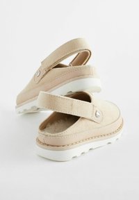 Chaussures en tissu beige avec un design à talon fermé, matériau texturé, intérieur doux et semelle en caoutchouc blanc avec un motif d'indentations.