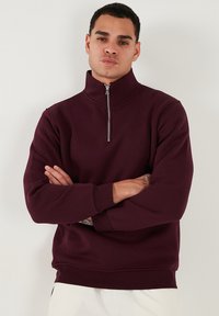 Sudadera burdeos con cremallera y cuello alto, puños y dobladillo acanalados. Tejido suave con una textura lisa y diseño minimalista.