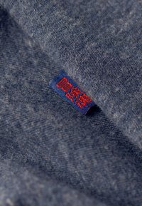 Tessuto blu-grigio con una superficie morbida e testurizzata. Un'etichetta blu con testo rosso è visibile, posizionata sul bordo del tessuto.