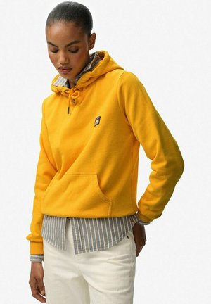 Sudadera con capucha amarilla de forro polar con diseño tipo pullover, bolsillo frontal y cordón ajustable. Capa con una camiseta de rayas azul y blanca por debajo.