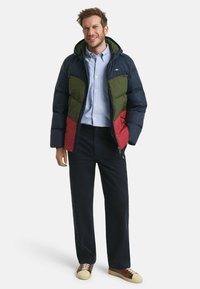 Gepolsterte Jacke mit Navy-, Grün- und Rotfarbblöcken, mit Kapuze, Reißverschluss und getragen über einem hellblauen Hemd und dunklen Hosen.