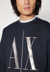 Felpa blu navy con grande logo "AX" argento sulla parte anteriore, colletto a costine e tessuto testurizzato. Dettaglio della parte superiore del torso.