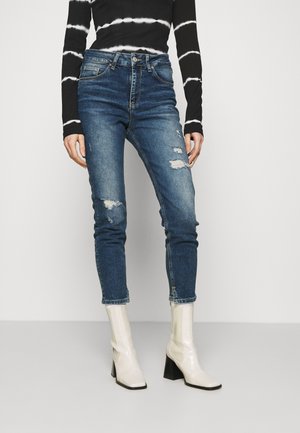 Vaqueros desgastados de color azul oscuro con un ajuste slim, que presentan desgarros y dobladillo deshilachado. Combinados con botas de tobillo blancas y brillantes con tacones de bloque.