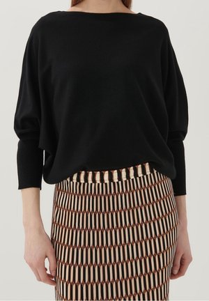 Femme portant un haut noir à manches longues col bateau rentré dans une jupe taille haute à motif géométrique rayé vertical.