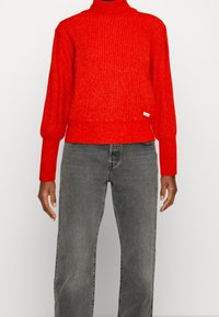 Persoon draagt een felrode geribbelde coltrui en vervaagde grijze rechte spijkerbroek tegen een effen achtergrond.