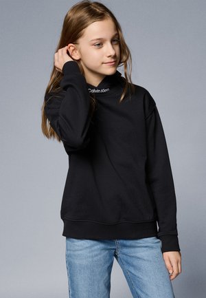 Juoda sportinė palaidinė su aukštu kaklu, raukšlėtais rankogaliais ir apatinėmis kraštinėmis. Ant apykaklės matomas baltas "Calvin Klein" logotipas. Dėvima su šviesiai mėlynomis džinsomis.