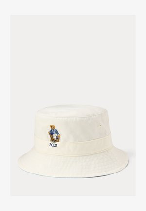 Off-white spandhat med broderet bjørn iført en blå blazer, siddende på en stol, med teksten "POLO" under bjørnen.