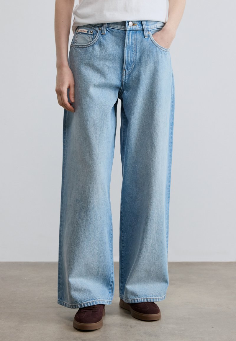 Φαρδιά τζιν παντελόνια σε ανοιχτό μπλε από denim με ψηλή μέση, πέντε τσέπες και διακριτική ξεθωριασμένη υφή, που συνδυάζονται με καφέ αθλητικά παπούτσια.