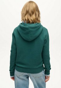Person med skulderlangt blondt hår iført en mørkegrøn hættetrøje og lyseblå jeans, vendt væk mod en simpel hvid baggrund.