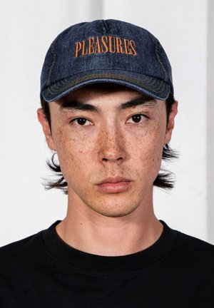 Młoda osoba z piegami, ubrana w ciemną jeansową czapkę z haftowanym na niej pomarańczowym napisem "PLEASURES" oraz czarną koszulkę na jednolitym tle.