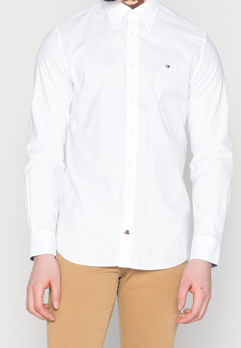 Homme portant une chemise blanche cintrée à manches longues boutonnée et un pantalon marron clair, debout les mains détendues le long du corps.
