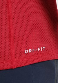 T-shirt de sport rouge perforé avec texte blanc « DRI-FIT » au-dessus d’un pantalon de sport bleu foncé avec coutures visibles.