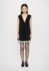 Μαύρο βελούδινο φόρεμα χωρίς μανίκια με βαθύ V-ντεκολτέ, διακοσμημένο με λευκές βούλες και κρόσσια στο τελείωμα. Το μοντέλο φοράει σκούρες καλσόν και παπούτσια.