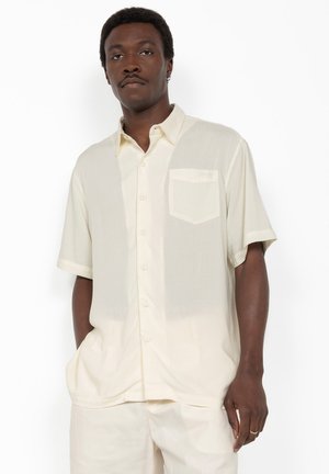 Homme portant une chemise crème à manches courtes avec une poche poitrine et un pantalon crème assorti, debout les mains dans les poches.