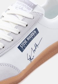 Λευκό αθλη τικό παπούτσι με ανοιχτό γκρι σουέντ λεπτομέρειες, λευκά κορδόνια, καουτσούκ σόλα, ετικέτα «Steve Madden EST 1990» και μαύρη υπογραφή στο πλάι.