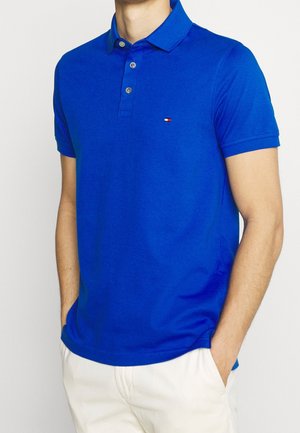 Polo en coton bleu avec un col, patte de boutonnage à trois boutons, manches courtes et un petit logo sur la poitrine. Texture lisse et design ajusté.