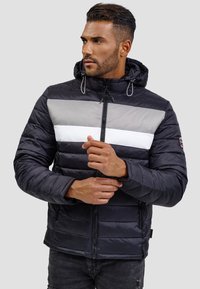 Chaqueta puffer negra con rayas horizontales grises y blancas, cierre con cremallera y capucha. Presenta una textura acolchada y bolsillos laterales.