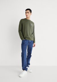 Zaļš garām piedurknēm krekls ar priekšējo grafiku. Apvienots ar ziliem džinsiem un baltām sporta apaviem ikdienišķam izskatam.