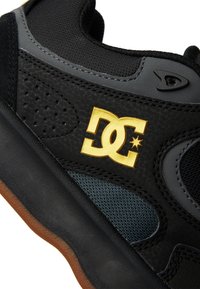 Black sneaker with textured black leather, gray mesh accents, and gum sole. Features a gold DC logo prominently displayed.
