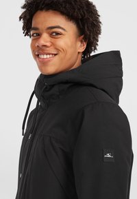 Leende person med kort lockigt hår iförd en svart huva-jacka med O'Neill-logotyp på ärmen mot en enfärgad bakgrund.