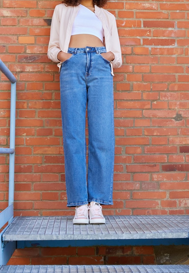 Světle růžová bomber bunda, bílý crop top, modré džíny s vysokým pasem a bílé tenisky na kovovém schodišti proti cihlové zdi.