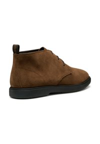 Botas de ante marrón hasta el tobillo con una suela de goma negra de perfil bajo, punta redonda y cordones mínimos. Cuenta con una lengüeta en el talón.