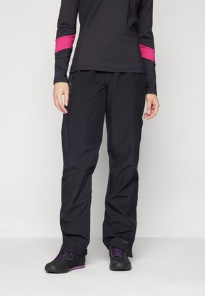Persoon draagt zwarte outdoorbroek, zwart shirt met lange mouwen met roze elleboogdetails en zwarte schoenen met paarse veters, staand tegen een witte achtergrond.