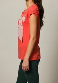 Koralowa koszulka krótkie rękawy z textured materiału i różową kokardą w kropki z przodu, w komplecie z zielonymi spodniami.